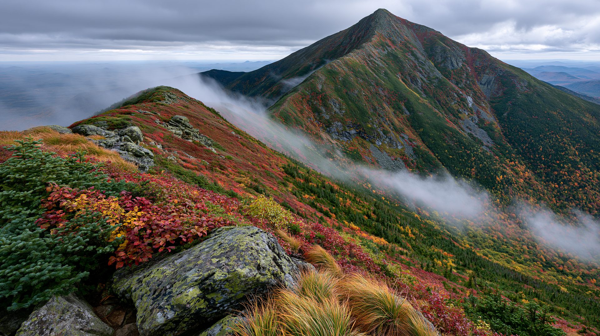A high-angle view of a rugged mountain ridge covered in red and yellow autumn tundra vegetation, with a thin line of white clouds drifting over the saddle toward a sharp green peak under an overcast sky.