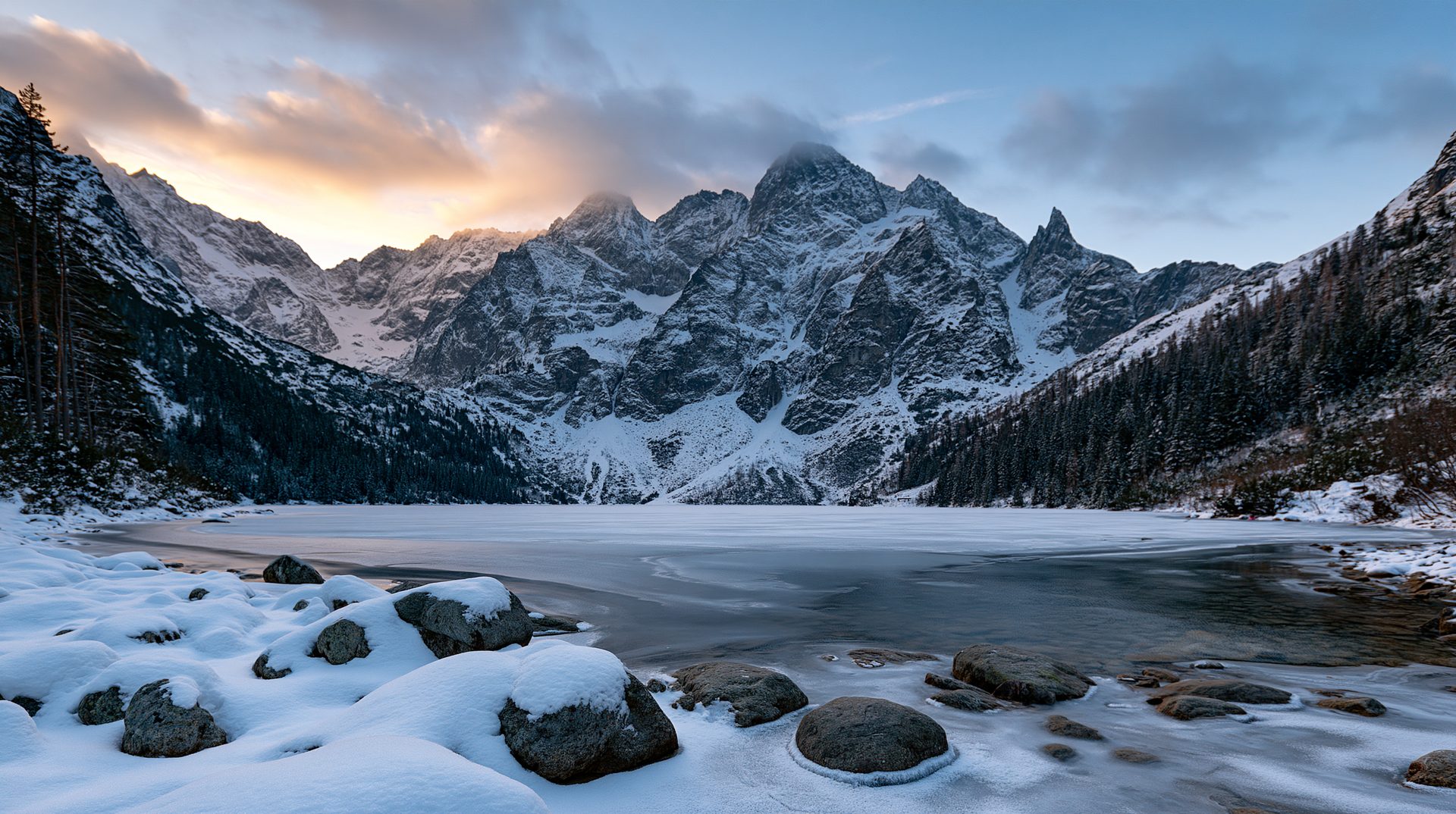A low-angle, wide-angle landscape photograph of a frozen alpine lake completely surrounded by snow-covered granite peaks under a golden winter sunset.