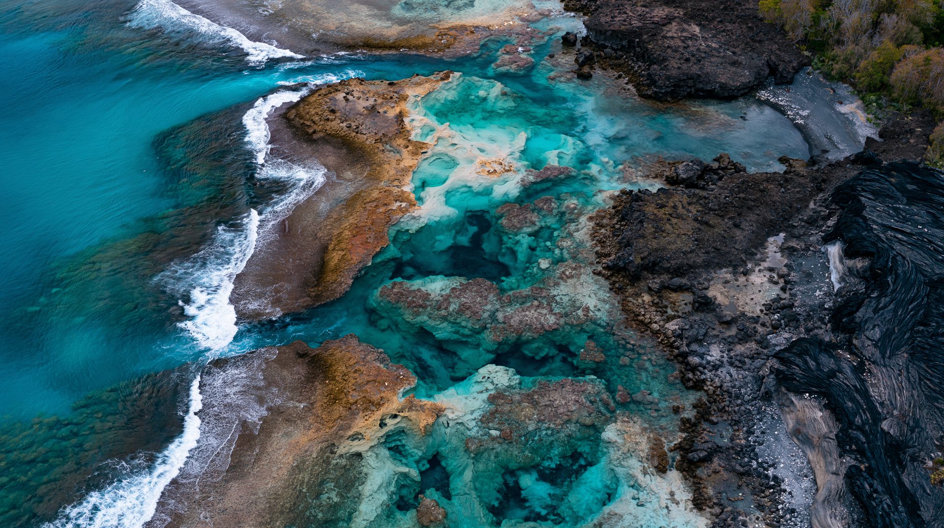 A high-angle aerial view of a jagged black volcanic rock coastline in Vanuatu, filled with bright turquoise tidal pools, channels, and crashing ocean waves breaking into white foam.