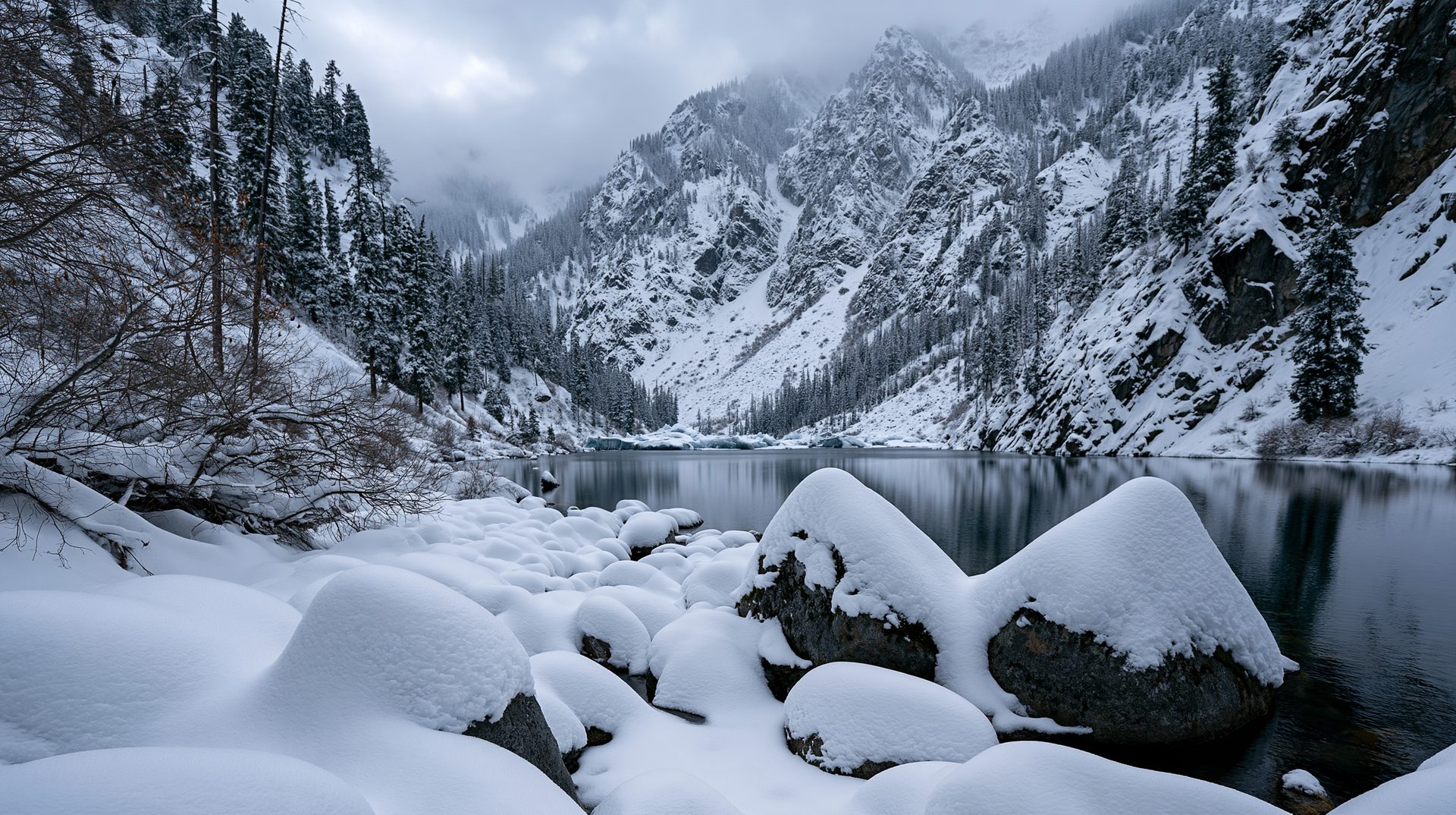 Smooth, heavy mounds of fresh snow cover large boulders along the edge of a calm, dark alpine lake, which reflects steep, snow-dusted mountain slopes and evergreen forests cloaked in low winter clouds.