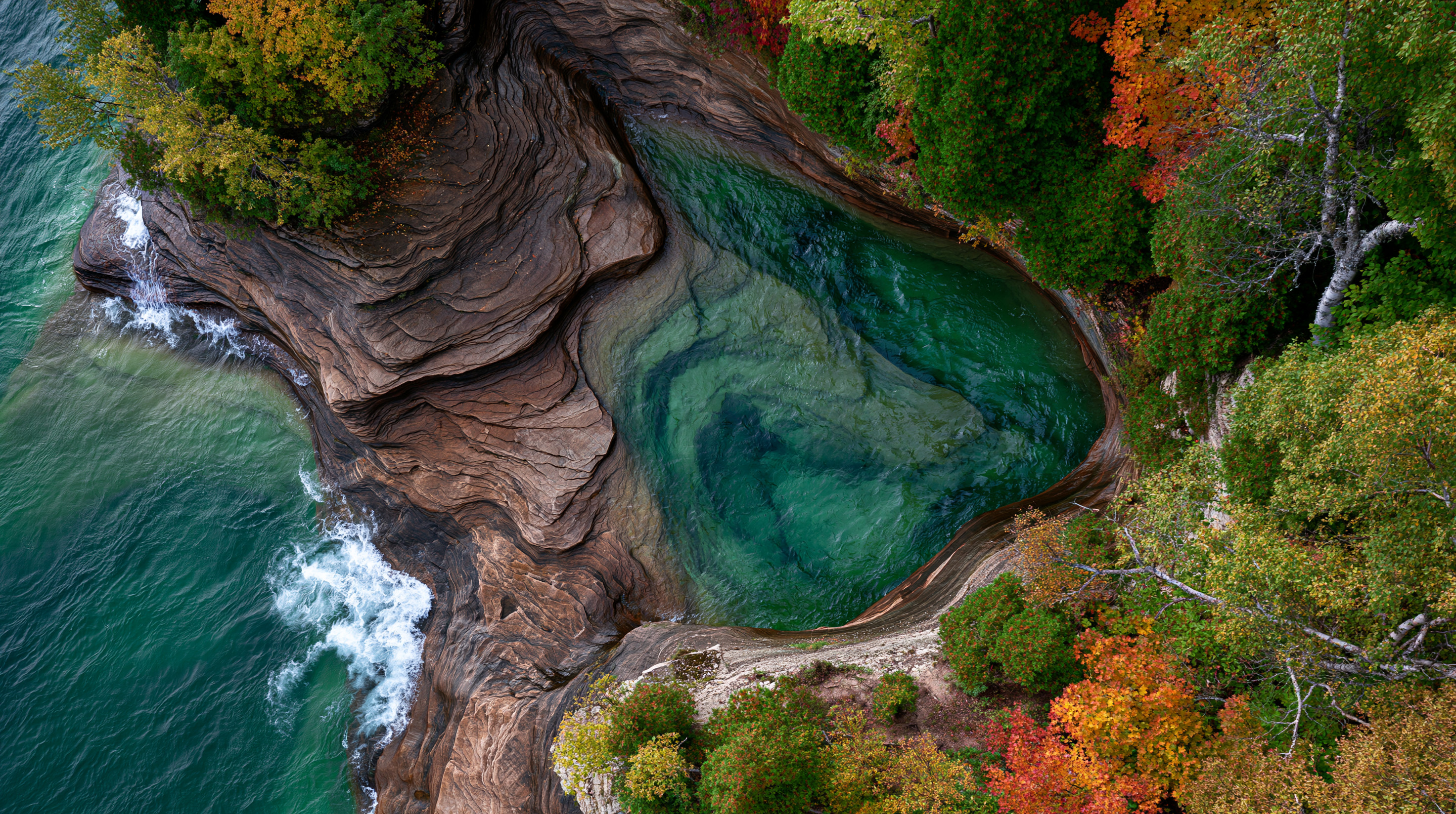 Top-down aerial drone photograph of a circular sandstone sea cave basin filled with clear green water, surrounded by rocky cliffs and autumn foliage on Lake Superior.