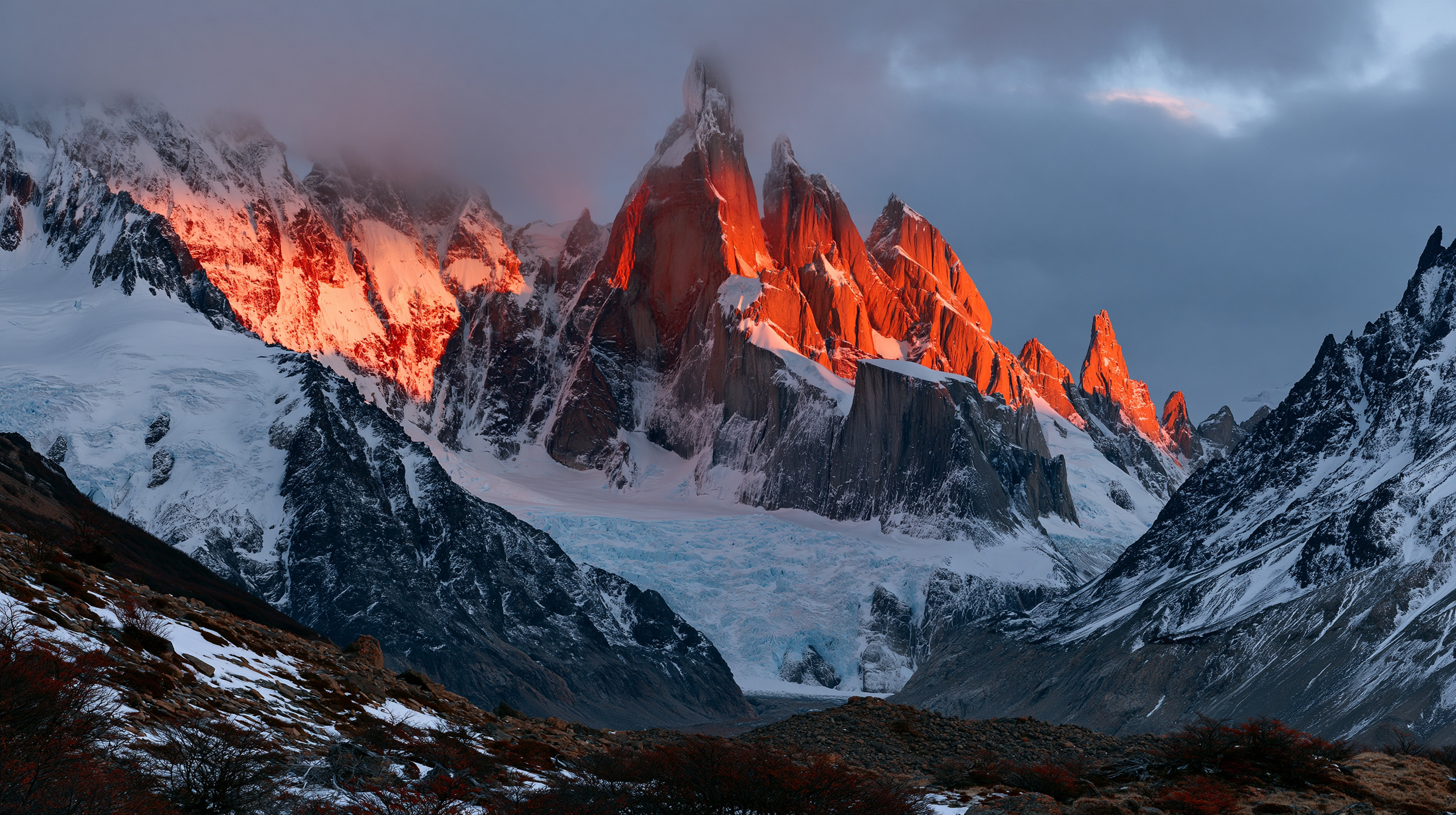 The sheer, jagged granite spires of Cerro Torre pierce a heavy blanket of storm clouds, their vertical rock faces set ablaze by a brilliant crimson sunrise, rising above a massive, fractured blue glacier carving through a snowy Patagonian mountain valley.