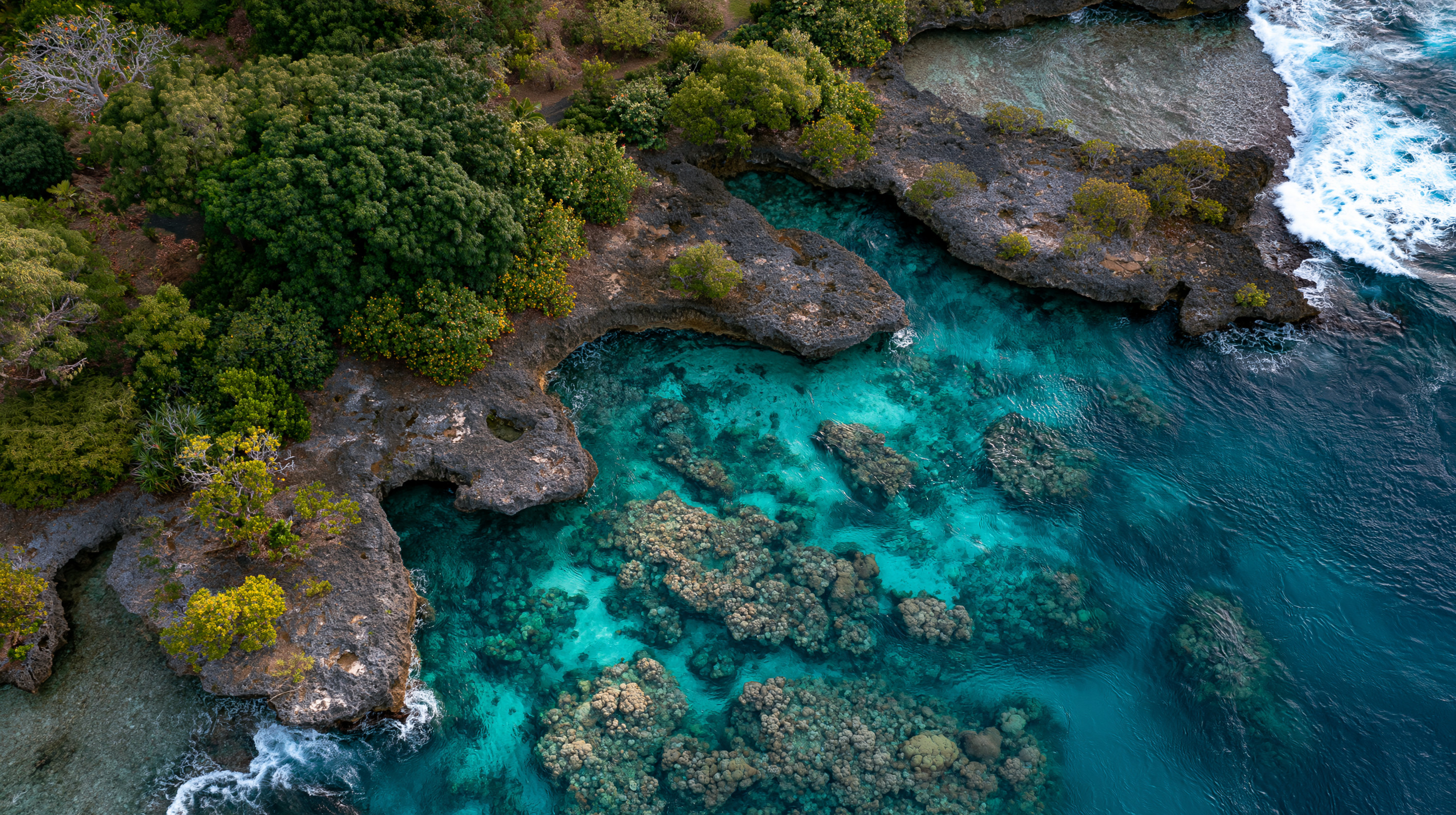 A high-angle aerial view of a rugged, dark limestone coastline in Vanuatu, enclosing a vibrant turquoise tidal pool with visible coral heads next to a deep blue sea with crashing white waves.