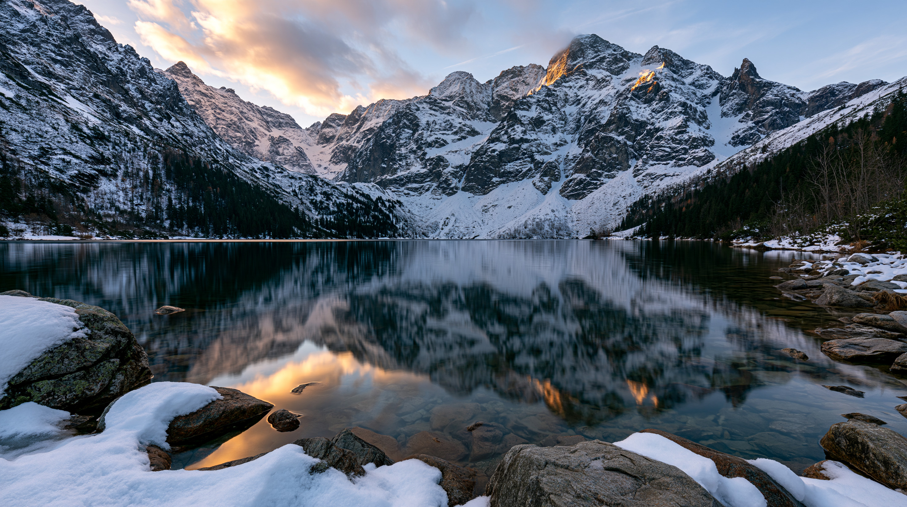 A wide-angle, symmetrical photograph of a mountain lake reflecting jagged, snow-capped granite peaks during a golden winter sunset.