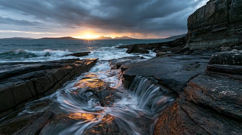 A dramatic wide-angle landscape photograph of ocean water cascading through dark, wet rocky channels along a Scottish coastline at sunset, featuring a bright sunburst breaking through heavy storm clouds.